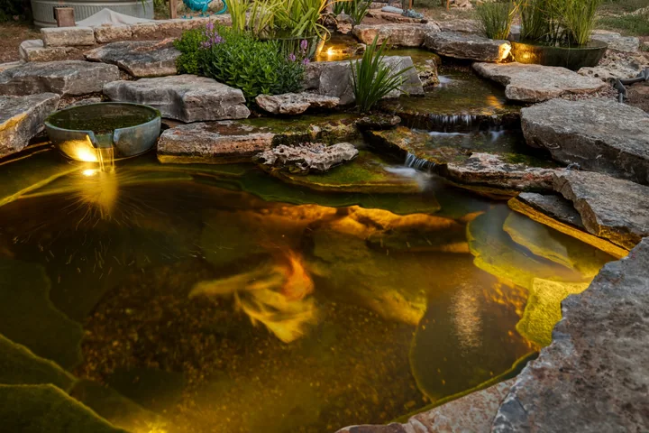 A koi pond glowing amber from underwater lighting beneath stone waterfalls at dusk
