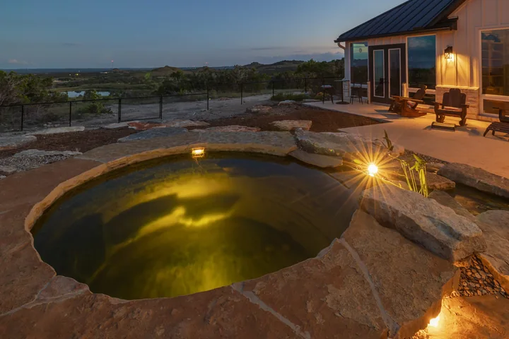 A circular stone-rimmed plunge pool glowing with underwater lighting beside a house at dusk