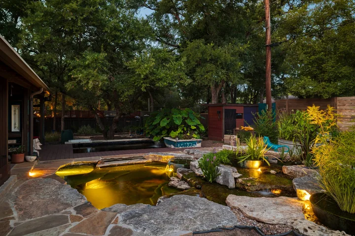 A warmly lit pond with stonework and planting beside a house at dusk